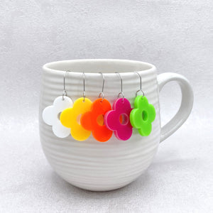 Colorful flower-shaped earrings hanging from a white mug on a light gray background