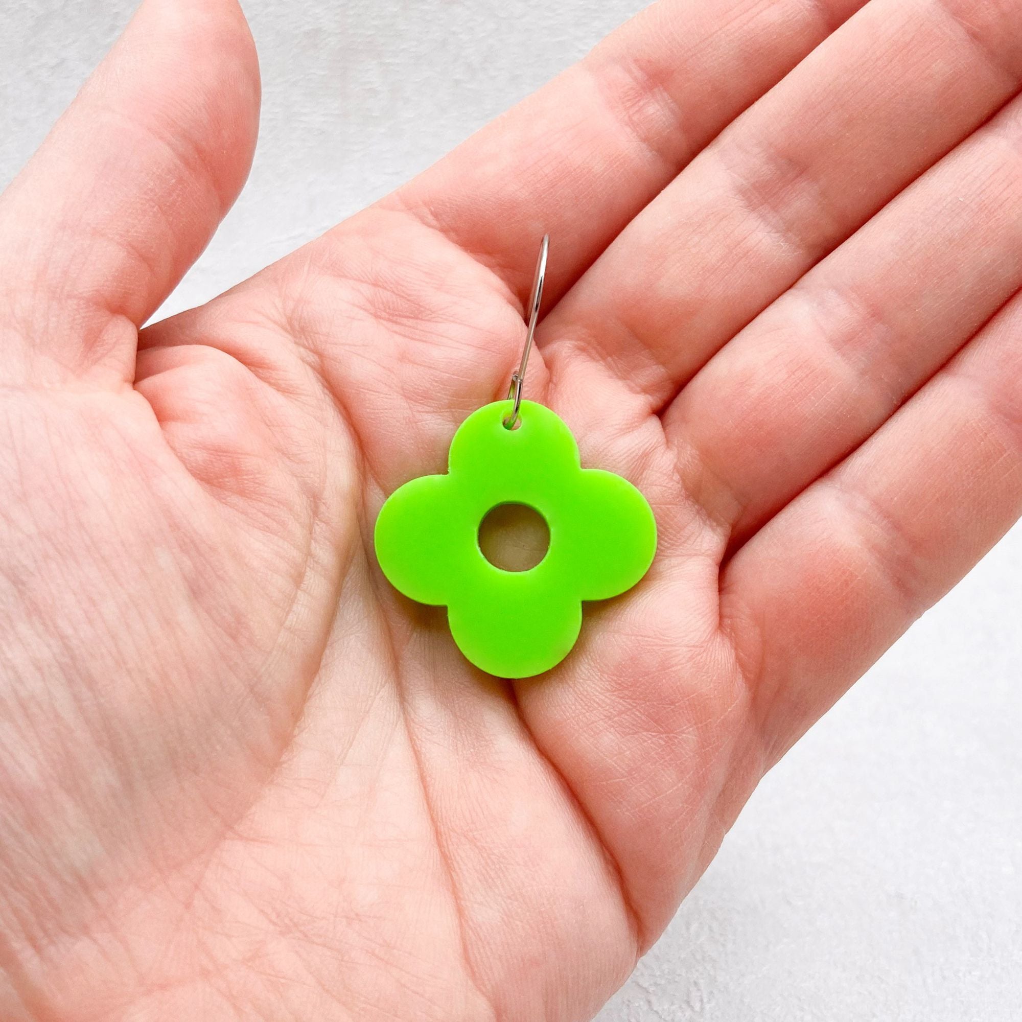 Hand holding green acrylic flower earring with four petals on a light grey background. 