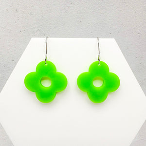 Four petal flower-shaped green acrylic earrings on a white hexagonal surface on a light grey background.