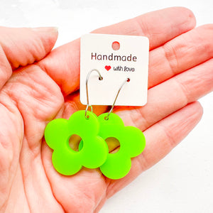 Hand holding green acrylic flower-shaped earrings with four petals and attached to a white card with "Handmade with Love' text on a light background.