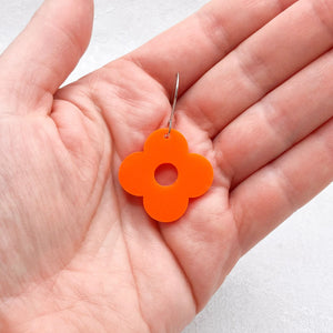 Hand holding an orange flower-shaped earring with four petals on a light grey background.