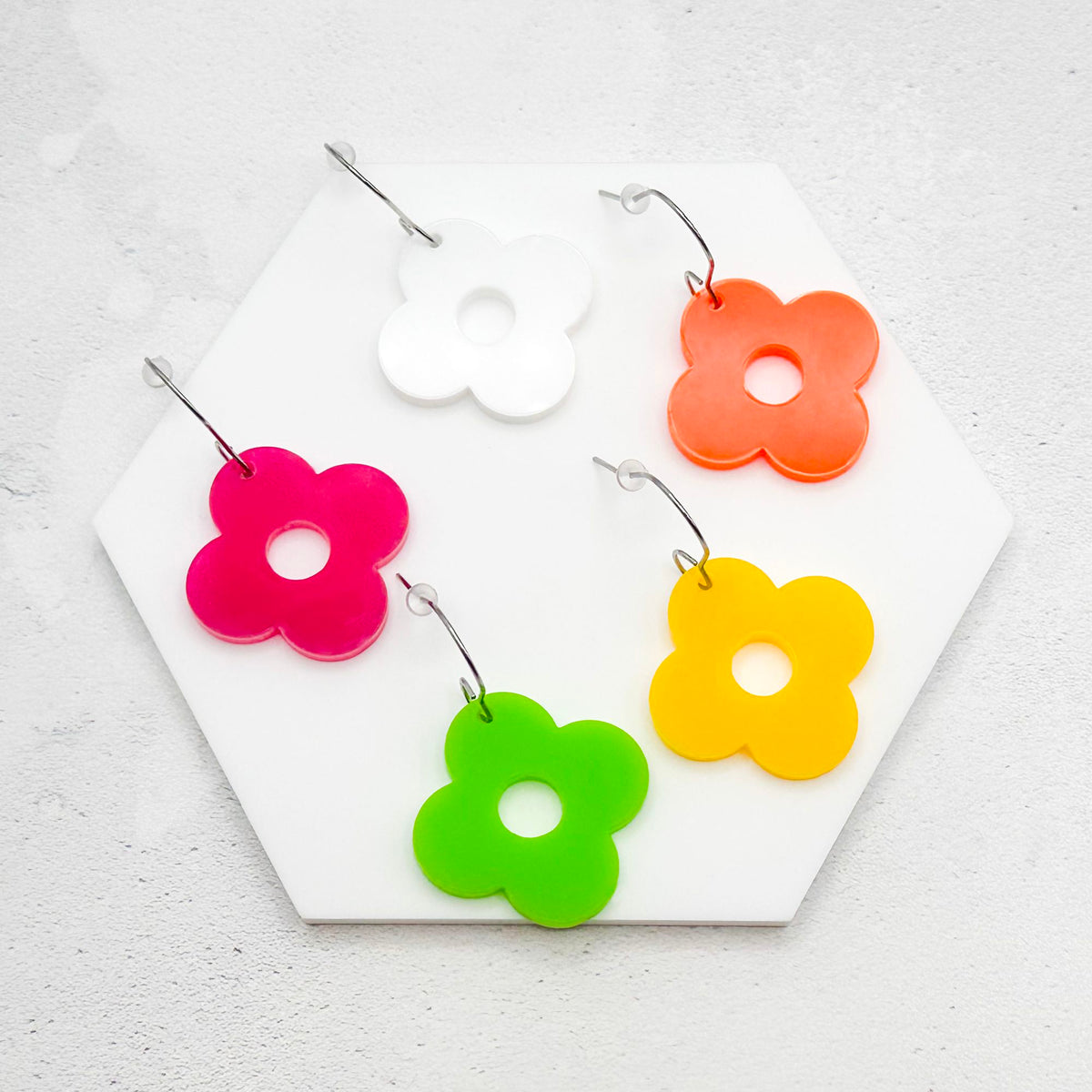 Acrylic flower earrings with four petals in multiple colours - white, orange, yellow, green, and pink, on a white marble surface on a light grey background.
