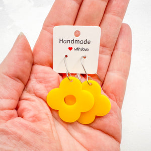 Hand holding yellow flower-shaped earrings attached to a white card with 'Handmade with Love' text on a light background.