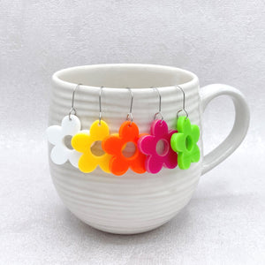 multiple coloured flower earrings suspended on a white ceramic mug on a grey background