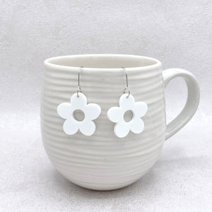 White flower-shaped earrings hanging from a white mug on a light gray background