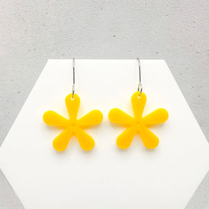 Yellow flower-shaped earrings on a white hexagonal stand against a light gray background