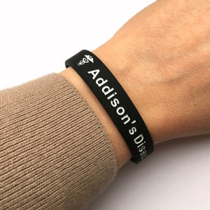 Black wristband with 'Addison's Disease' text on a wrist wearing a beige sleeve.