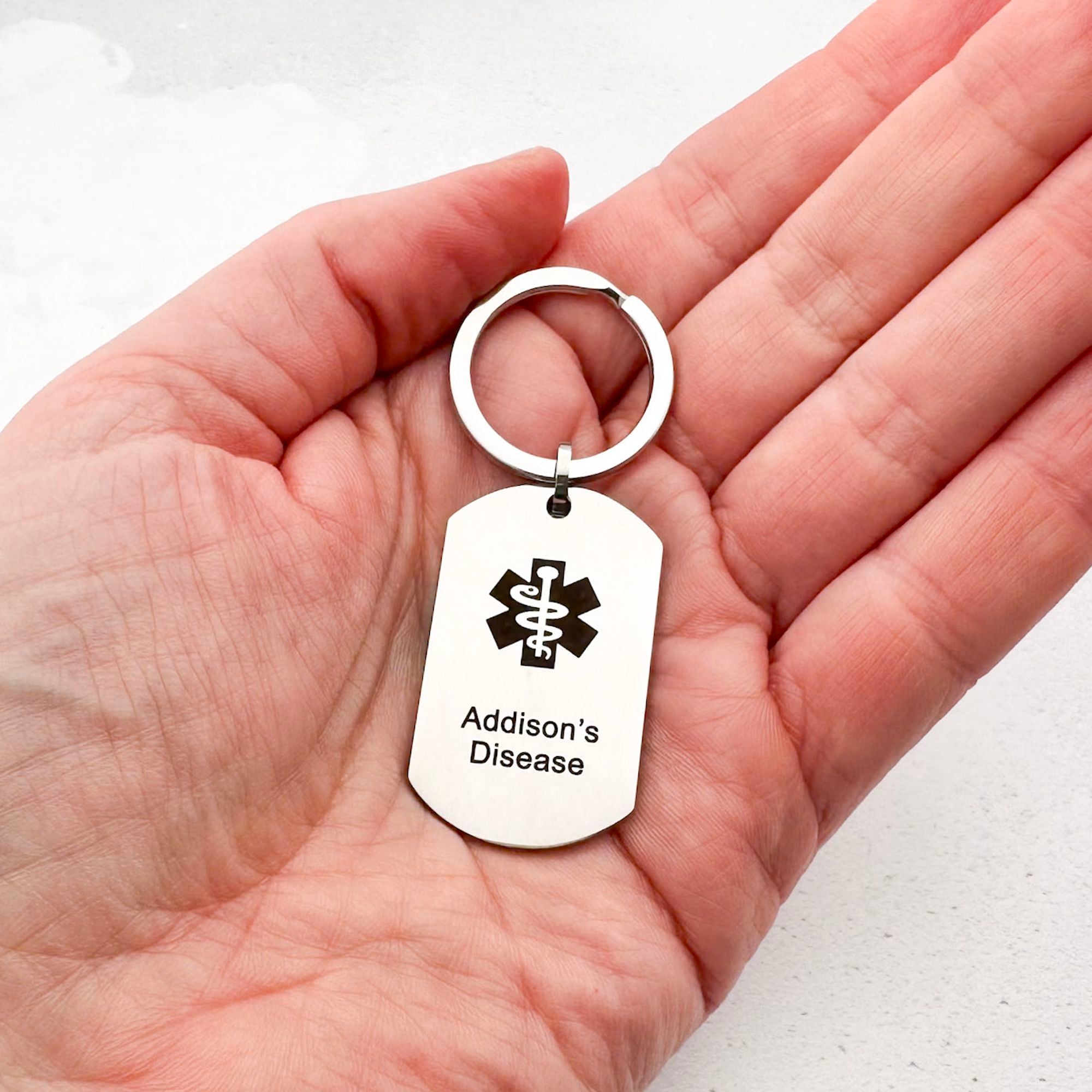 Hand holding a stainless steel keychain with Addison's Disease text on a white background