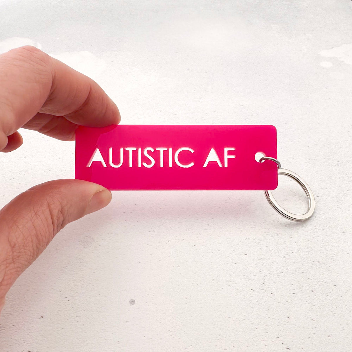 Pink keychain with 'AUTISTIC AF' text held by a hand on a white background
