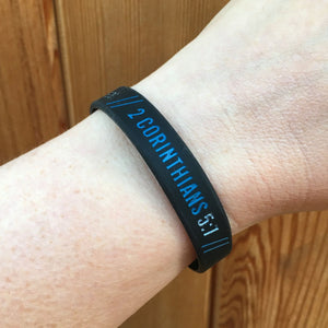 Black wristband with '2 Corinthians 5:7' text on a wrist against a wooden background