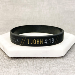 Black wristband with '1 John 4:19' text on a white stand