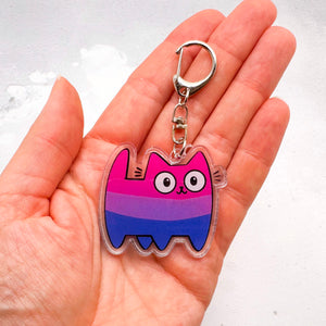 A hand holding a pink, purple, and blue bisexual striped cat-shaped acrylic keychain with a silver clasp.