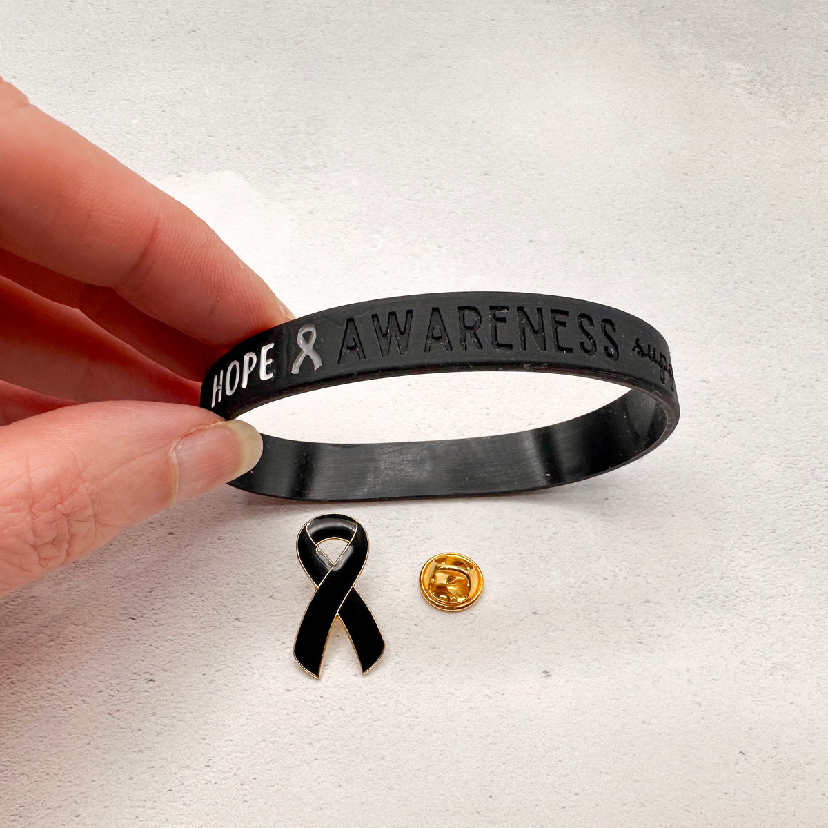 Black 'Hope & Awareness' bracelet held by a hand with a black ribbon and gold button on a light gray background.