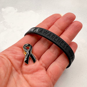 black silicone wristband engraved with ‘Hope & Awareness’ held in a person's hand with matching black enamel ribbon pin