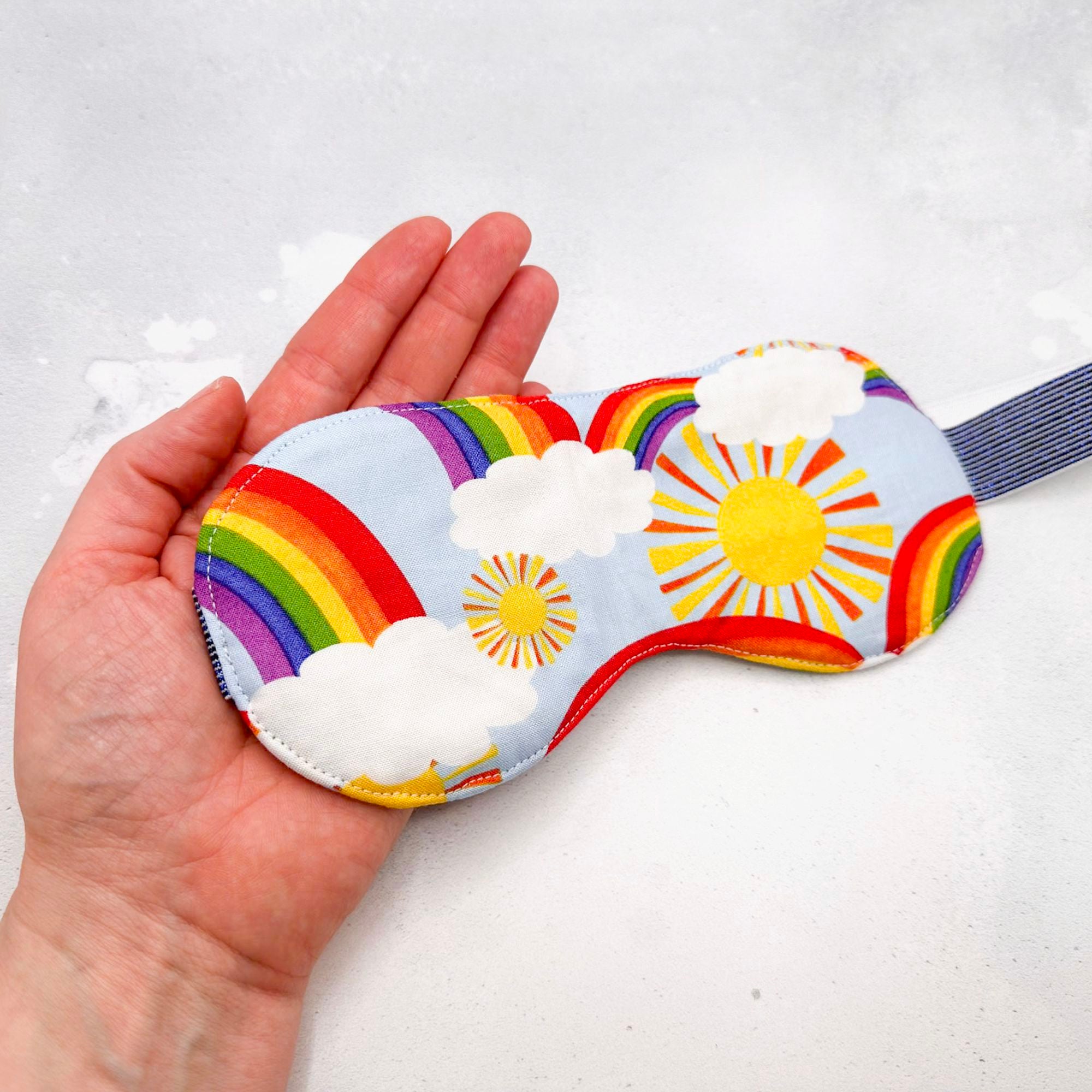 Hand holding a colourful sleep mask with rainbow, sun, and cloud design 