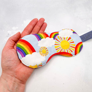 Hand holding a colourful sleep mask with rainbow, sun, and cloud design 