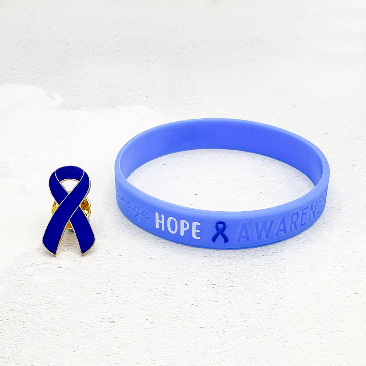 blue silicone awareness ribbon wristband with "Hope & Awareness" text engraved on its side, and a blue enamel ribbon pin besides it
