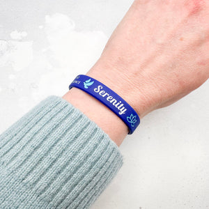 A blue silicone wristband with the word 'Serenity' and small white icons, worn on a person's wrist.