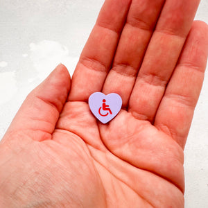 purple acrylic heart pin with disabled symbol in red held in a hand for size