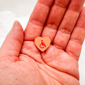 orange acrylic heart pin with disabled symbol in red held in hand
