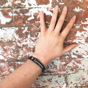 Hand wearing a black wristband with 'Epilepsy' text against a textured brick wall.