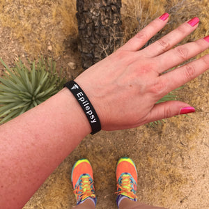 Hand wearing a black 'Epilepsy' wristband with colourful sneakers on a natural background