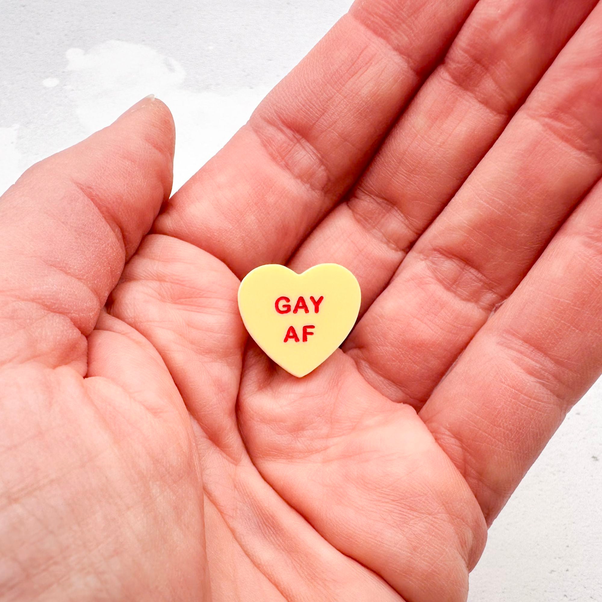 yellow acrylic heart pin with "Gay AF" hand-painted in red held in hand