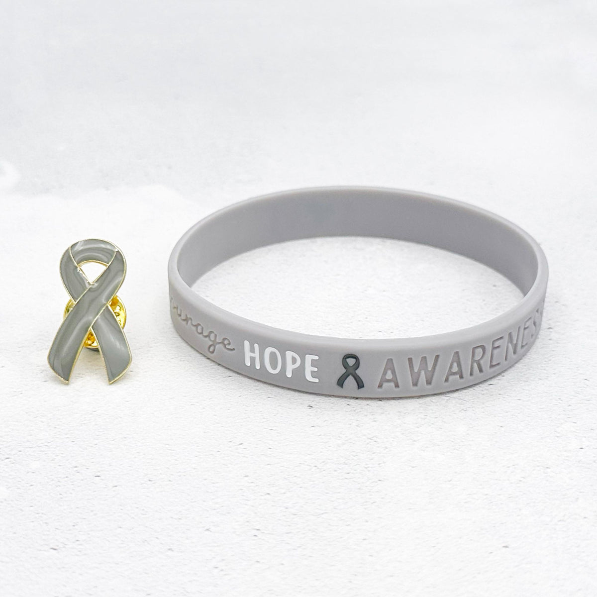 grey awareness ribbon wristband with "hope & awareness" text engraved on its side and a matching grey enamel ribbon pin next to it