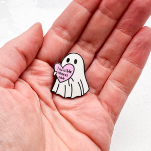 Ghost-shaped pin with a pink heart labelled 'Invisible Illness Club' held in a hand on a white background