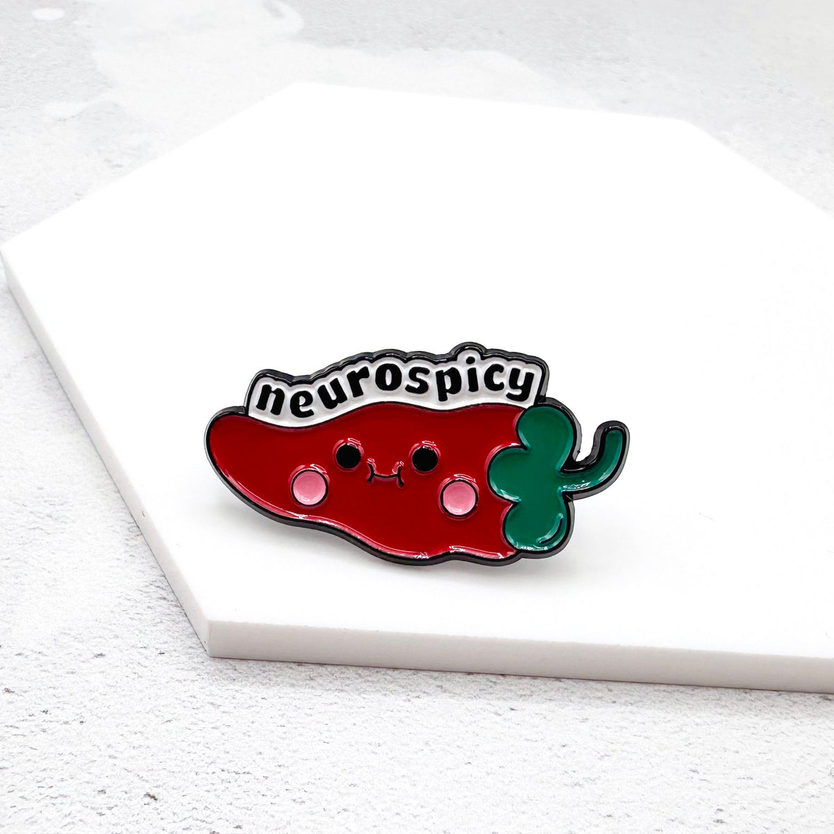 Enamel pin with a red and green pepper design labelled 'neurospicy' on a white surface.