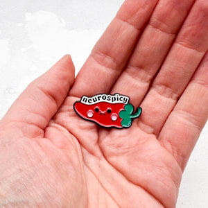 Hand holding a red and green enamel chilli pepper pin with 'neurospicy' text on a white background