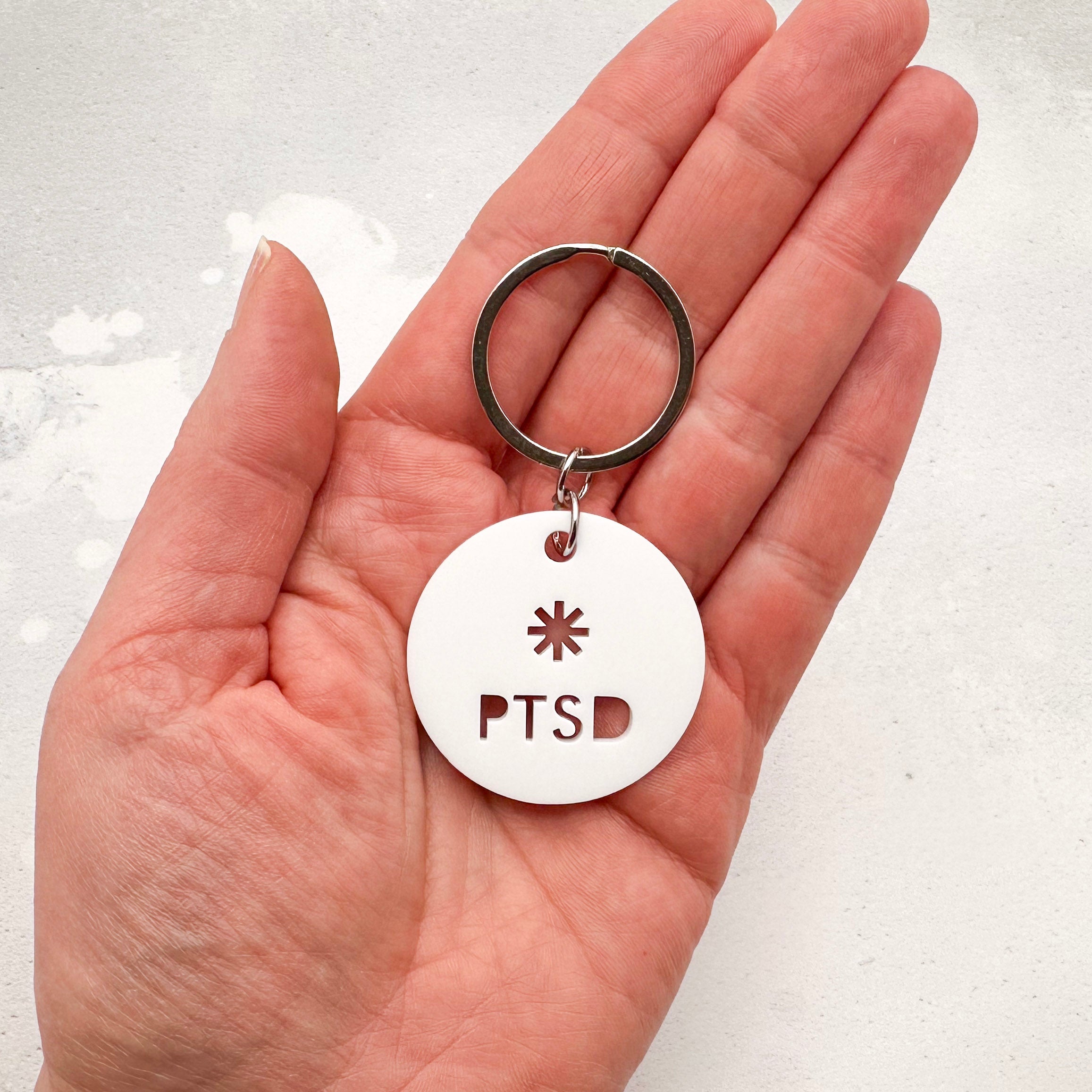 lightweight white acrylic round keychain with 'PTSD' laser cut out, held in hand