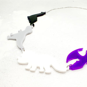 Necklace with four dinosaur-shaped pendants in the asexual colours of black, grey, white, and purple, on a light grey background.