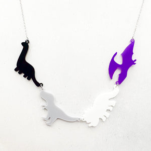 Necklace with four dinosaur-shaped pendants in the asexual colours of black, grey, white, and purple on a light grey background.