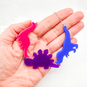 Hand holding necklace with three dinosaur-shaped pendants in bold bisexual colours of pink, purple, and blue, on a light grey background.