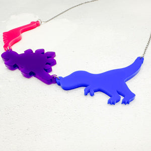 Necklace with three dinosaur-shaped acrylic pendants in bold bisexual colours of pink, purple, and blue, on a light grey background. 