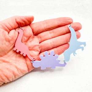 Hand holding necklace with three dinosaur-shaped pendants in pastel bisexual colours of pink, purple, and blue, on a light background.