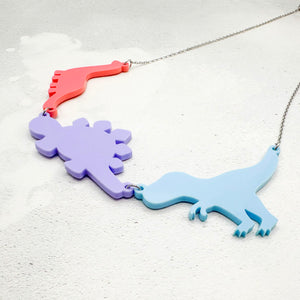 Necklace with three dinosaur-shaped pendants in pastel bisexual colours of pink, purple, and blue, on a light grey background.