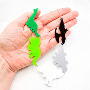 Hand holding necklace with five dinosaur-shaped pendants in the aromantic colours of green, light green, white, grey, and black, on a light background.
