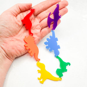 Hand holding a necklace with six dinosaur-shaped pendants in the gay rainbow colours of red, orange, yellow, green, blue, and purple, on a light background.