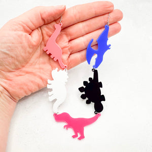 Hand holding necklace with dinosaur-shaped pendants in the genderfluid colours of pink, white, hot pink, black, and blue, on a light background.