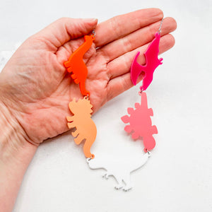 Hand holding a necklace with five dinosaur-shaped pendants in the lesbian colours of orange, white, and pink, on a white background