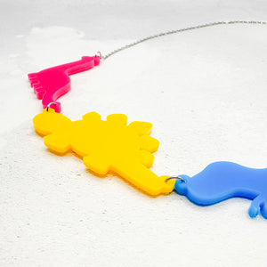 Necklace with three dinosaur-shaped pendants in the pansexual colours of pink, yellow, and blue, on a light background.