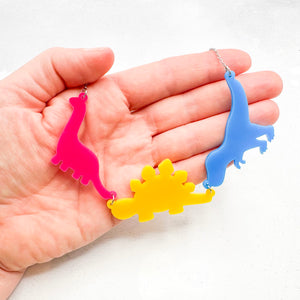 Hand holding a necklace with three dinosaur-shaped pendants in the pansexual colours of pink, yellow, and blue, on a light background.