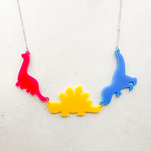 Necklace with three dinosaur-shaped pendants in the pansexual colours of pink, yellow, and blue, on a light grey background.