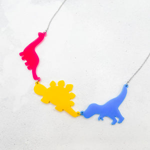 Necklace with three dinosaur-shaped pendants in the pansexual colours of pink, yellow, and blue, on a light grey background