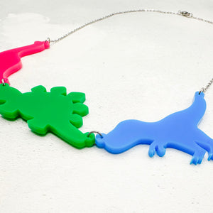 Necklace with three dinosaur-shaped pendants in the polysexual colours of pink, green, and blue on a textured light grey background