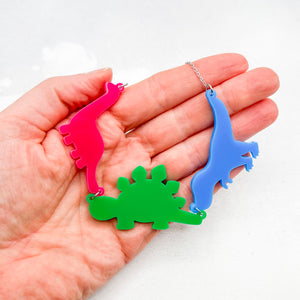 Hand holding necklace with three dinosaur-shaped pendants in the polysexual colours of pink, green, and blue, on a light grey background