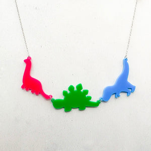 Necklace with three dinosaur-shaped pendants in the polysexual colours of pink, green, and blue, on a light grey textured background