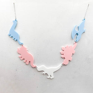 Necklace with five dinosaur-shaped pendants in pastel transgender colours of blue, pink, and white, on a light grey background.